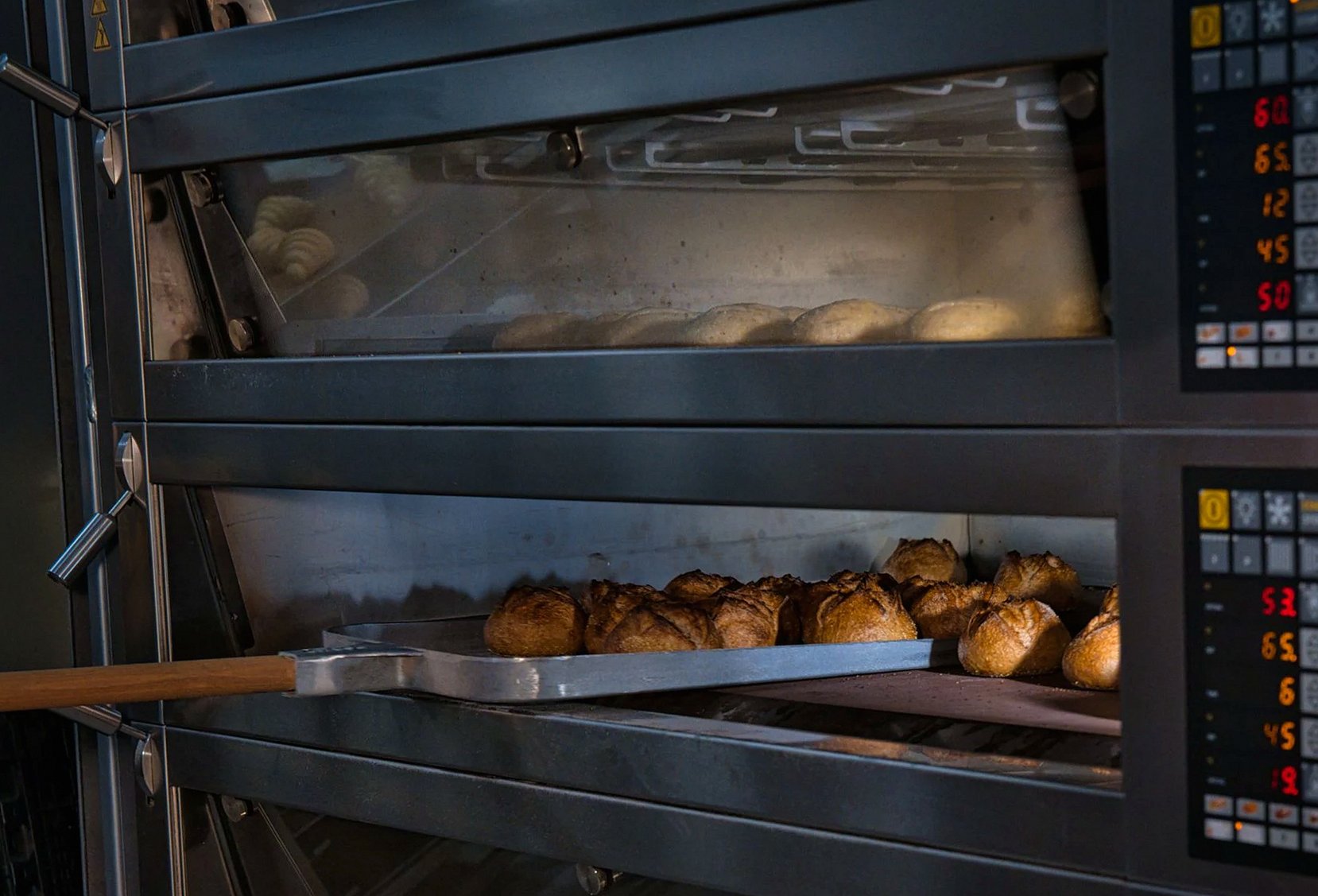 Golden-brown sourdough loaves on a tray being pulled from the lower deck of a deck oven during the morning bake.